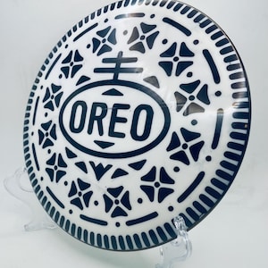 Könnte beinhalten: Eine weiß-schwarze dekorative Platte mit dem Wort "OREO" in der Mitte. Die Platte zeigt ein detailliertes Design aus schwarzen Formen und Mustern, das einem Oreo-Keks ähnelt. Die Platte wird auf einem transparenten Ständer ausgestellt.