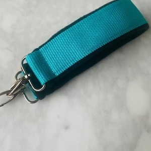 Puede incluir: Una correa de muñeca turquesa con un borde negro y un cierre de metal plateado. La correa está hecha de tela tejida. El fondo es una superficie veteada gris claro y blanco.