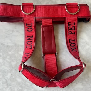 Peut inclure: Un harnais rouge doté de deux sangles verticales avec le texte brodé en noir DO NOT PET. Le harnais est fabriqué à partir d'une combinaison de sangle rouge et de matière rouge lisse, agrémenté d'anneaux en D et de boucles de réglage en métal argenté.