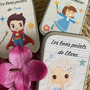 Peut inclure: Trois petites boîtes métalliques rectangulaires avec des illustrations de dessins animés et du texte. Une boîte présente un super-héros, une autre une fée et la troisième un personnage de type fée. Le texte sur les boîtes se lit "Les bons points de Tom", "Les bons points de Philomène" et "Les bons points de Clara".