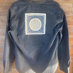 May include: A dark blue long-sleeve shirt with a square, decorative patch on the back. The patch features a white, crocheted doily design surrounded by a blue and brown patterned border. The shirt is button-down.