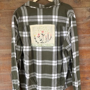May include: Green and white plaid flannel shirt with a floral embroidered patch on the back. The patch features pink flowers and green stems. The shirt is hanging on a white hanger.