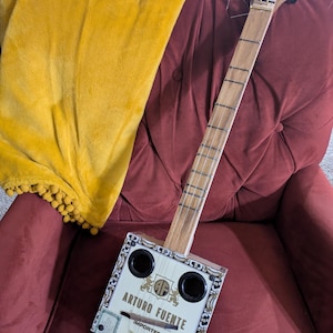 May include: A handmade cigar box guitar with a wooden neck and fretboard. The rectangular body is made from an "Arturo Fuente" cigar box with black circular sound holes. The guitar rests on a burgundy armchair with a yellow blanket.