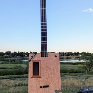 May include: A handmade cigar box bass guitar with a light brown wooden body and a long, black neck. The instrument has four tuning pegs and a small, rectangular sound hole. A small wooden box sits at the base of the guitar.