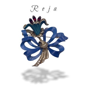 Pode incluir: Um broche de esmalte azul e strass com uma flor estilizada com um laço. A flor tem um centro turquesa e strass vermelhos.