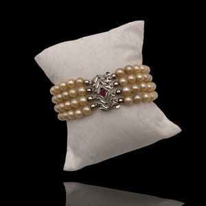 以下が含まれることがあります： 葉のデザインと小さな赤い宝石が付いたシルバーの留め具が付いた4連のパールブレスレットです。