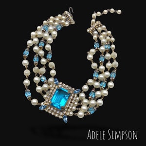 May include: A vintage necklace featuring multiple strands of faux pearls and light blue crystal accents. The focal point is a large, square, light blue crystal surrounded by smaller crystals. The necklace is labeled "ADELE SIMPSON".