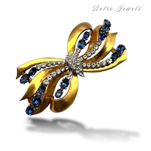 Pode incluir: Broche vintage dourado em forma de laço com detalhes em cristal azul e transparente. O broche apresenta um design de laço com um aglomerado central de cristais transparentes e é adornado com cristais azuis ovais. As palavras "Retro Jewels" estão no canto superior direito.