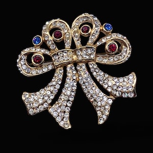 Puede incluir: Broche de lazo dorado incrustado con cristales transparentes. El diseño del lazo presenta piedras de acento rojas y azules. El broche está sobre un fondo negro, resaltando su brillo y detalles intrincados. Una pieza de joyería clásica.