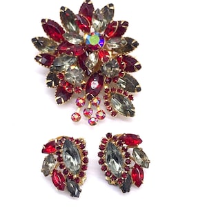 Peut inclure: Un ensemble de bijoux vintage comprenant une broche florale et des boucles d'oreilles assorties. La broche et les boucles d'oreilles sont ornées de strass rouges, gris et irisés sertis dans du métal doré. La broche a un design en cascade, et les boucles d'oreilles sont un groupe de pierres.