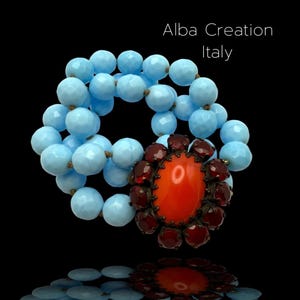 Può includere: Un braccialetto vintage con perline sfaccettate azzurre e un motivo floreale centrale arancione e bordeaux. Il braccialetto è di Alba Creation Italy. Il design è su sfondo nero.
