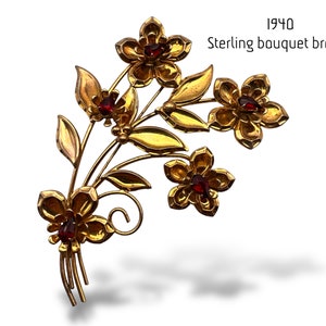 Könnte beinhalten: Eine goldfarbene Blumenstrauß-Brosche aus Metall mit vier fünfblättrigen Blüten, die tiefrote, tropfenförmige Zentren sowie schlanke Stiele und Blätter aufweisen. Der Text 1940 Sterling bouquet brooch ist oben rechts im Bild zu sehen.