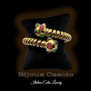 Puede incluir: Dos pulseras doradas en espiral con terminales con incrustaciones de joyas. Cada terminal presenta una gema roja central rodeada de gemas verdes y azules. Las pulseras se exhiben sobre un cojín de terciopelo negro.