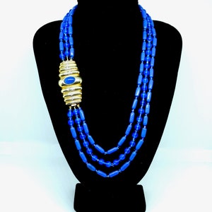 Op de afbeelding: Een meerstrengs ketting met drie rijen cilindrische blauwe kralen. De strengen zijn verbonden door een grote, goudkleurige metalen sluiting die horizontaal geribbeld is en is geaccentueerd met een centrale ovale blauwe steen en kleine heldere kristallen.