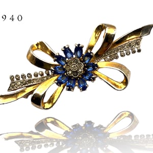 Pode incluir: Um broche dourado com um design de laço de cristal azul e transparente. O broche tem a forma de um laço com uma flor no centro. A flor é feita de cristais azuis e transparentes.
