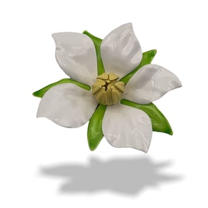 Könnte beinhalten: Eine weiße Blumenbrosche mit grünen Blättern und einer gelben Mitte. Die Blume ist aus Emaille und hat eine glänzende Oberfläche.