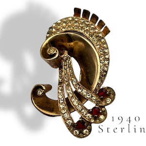 Puede incluir: Un broche dorado con un diseño estilizado que presenta una forma curva con una forma curva más pequeña en la parte superior. El broche está decorado con diamantes de imitación transparentes y piedras rojas. El texto "1940 Sterling" es visible debajo del broche.