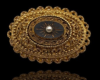 May include: A gold-toned brooch with an oval-shaped black enamel center featuring a starburst design with a pearl in the center. The brooch is surrounded by a decorative filigree border.