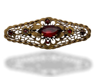 Op de afbeelding: Een gouden filigraan broche met een grote rode ovale edelsteen in het midden. De broche is versierd met kleinere rode edelstenen aan de randen.