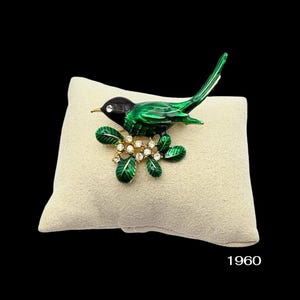 May include: A vintage bird brooch with a black head and vibrant green body, perched on a branch with crystal accents. The brooch is set against a beige background. The number "1960" is visible in the bottom right corner.