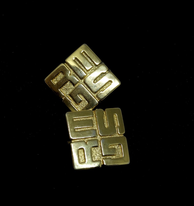May include: Two gold-colored metal square-shaped earrings with an intricate design of interlocking squares.
