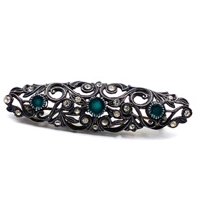 May include: An ornate silver brooch with a delicate filigree design. The brooch is decorated with clear rhinestones and three green gemstones.
