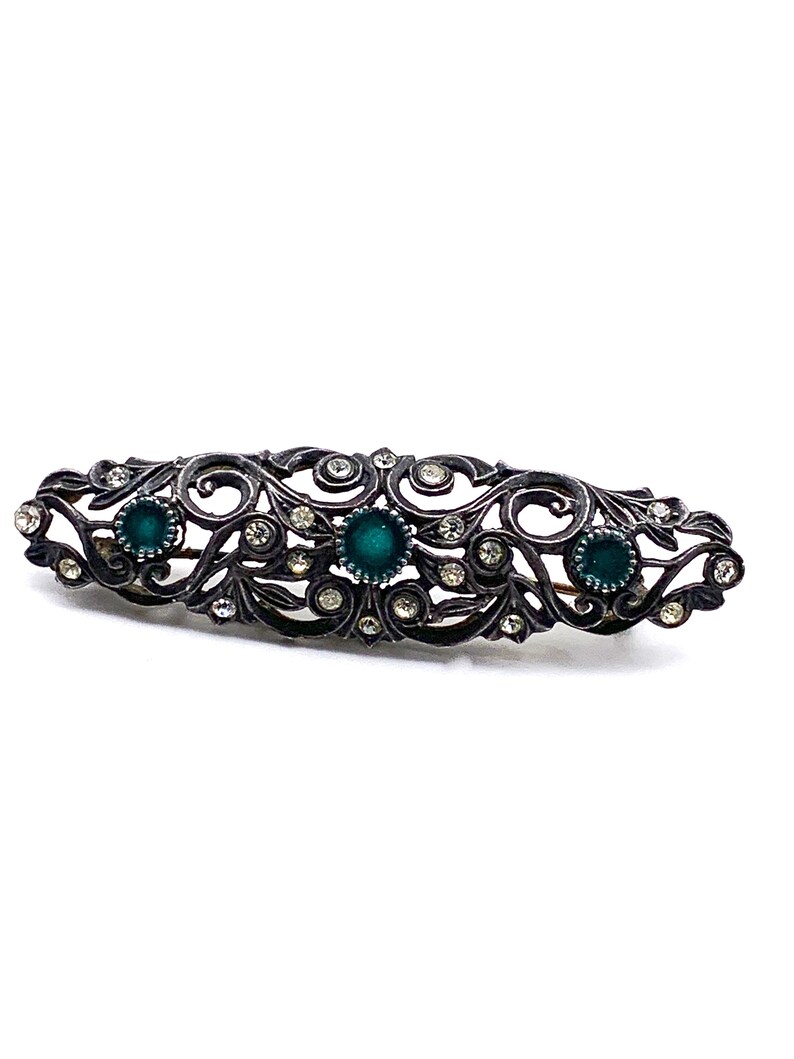May include: An ornate silver brooch with a delicate filigree design. The brooch is decorated with clear rhinestones and three green gemstones.