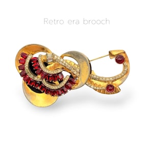 May include: A vintage brooch with a gold-tone metal base, featuring red and clear crystal accents. The brooch has a curved design with a pin closure and the text "Retro era brooch".