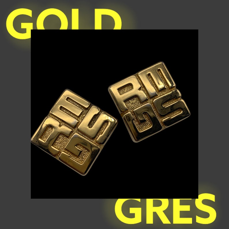 May include: A pair of gold-tone earrings with a geometric design. Each earring features a square shape with the letters "GRES" embossed on the surface.