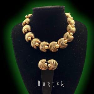 May include: A gold-toned necklace and matching earrings set. The necklace and earrings feature a seashell design with a pearl in the center of each shell. The jewelry is displayed on a black mannequin head. The word "Bartek" is written in white.