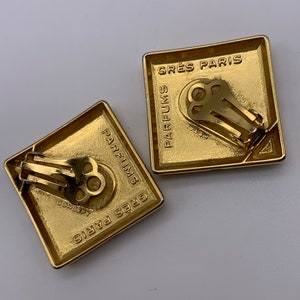 May include: A pair of gold-tone clip-on earrings with a square shape. The earrings feature the text "GRES PARIS PARFUMS" on the front and back.
