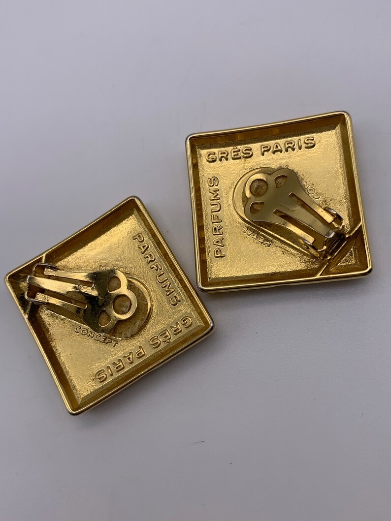 May include: A pair of gold-tone clip-on earrings with a square shape. The earrings feature the text "GRES PARIS PARFUMS" on the front and back.
