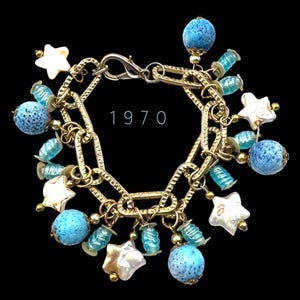 Könnte beinhalten: Ein goldenes Kettenarmband mit mehreren Anhängern, darunter blaue und weiße sternförmige Anhänger, blaue Lavasteine und blaue Glasperlen. Das Armband hat einen Verschluss und die Zahl "1970" ist auf der Kette sichtbar.