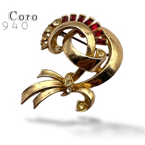 Könnte beinhalten: Goldfarbene Coro Brosche mit roten und klaren Strasssteinen. Die Brosche ist in Form einer stilisierten Schleife mit einem Wirbelmuster gestaltet. Die Brosche ist mit "Coro 1940" gekennzeichnet.