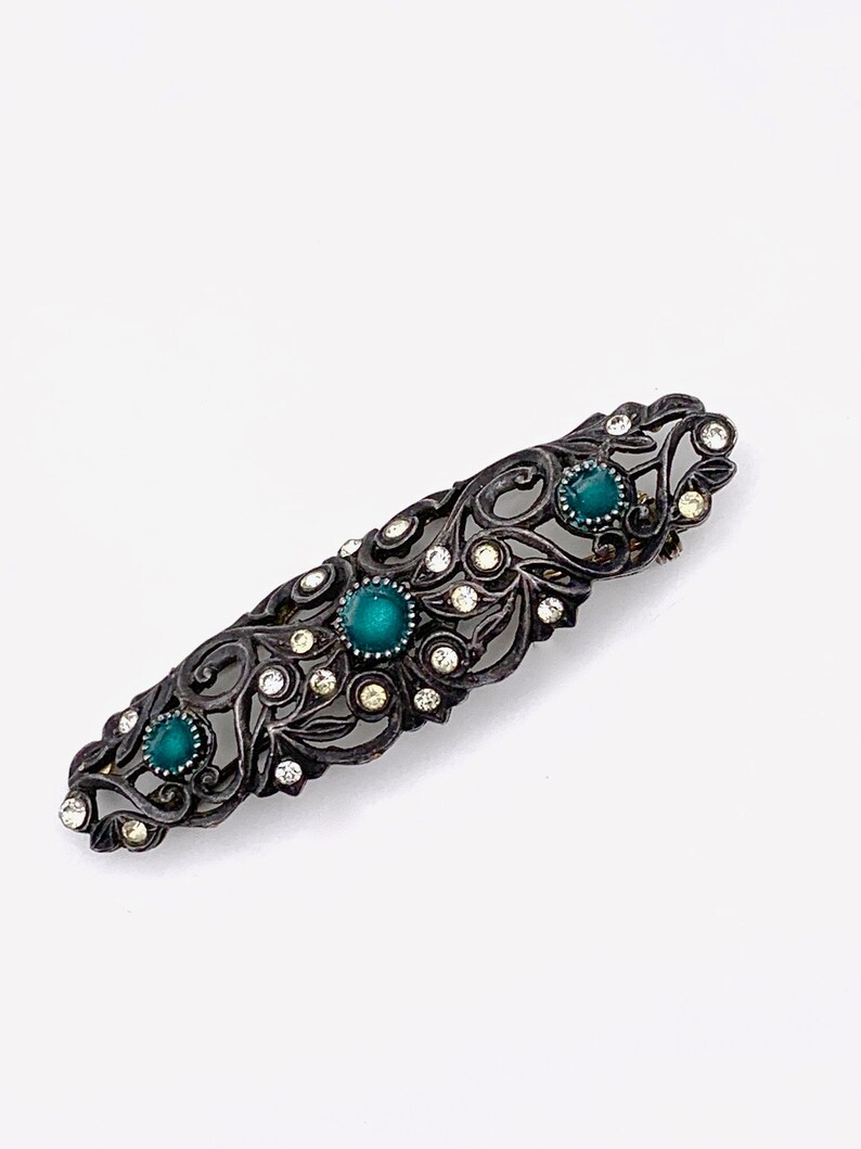 May include: A vintage brooch with a filigree design featuring a dark metal base and green and clear rhinestones.