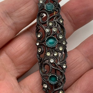 May include: A silver-toned hair clip with a floral design and three green stones. The clip is decorated with clear rhinestones.