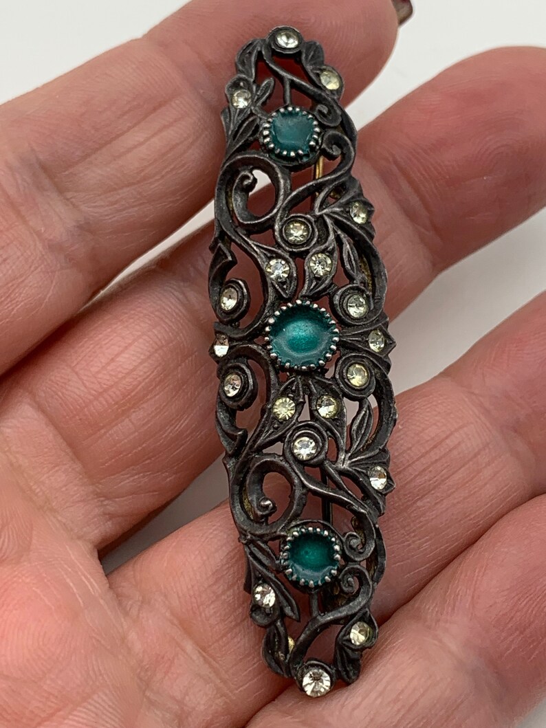 May include: A silver-toned hair clip with a floral design and three green stones. The clip is decorated with clear rhinestones.