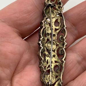 May include: A silver filigree brooch with a gold tone finish and a pin closure. The brooch is decorated with small, round stones.