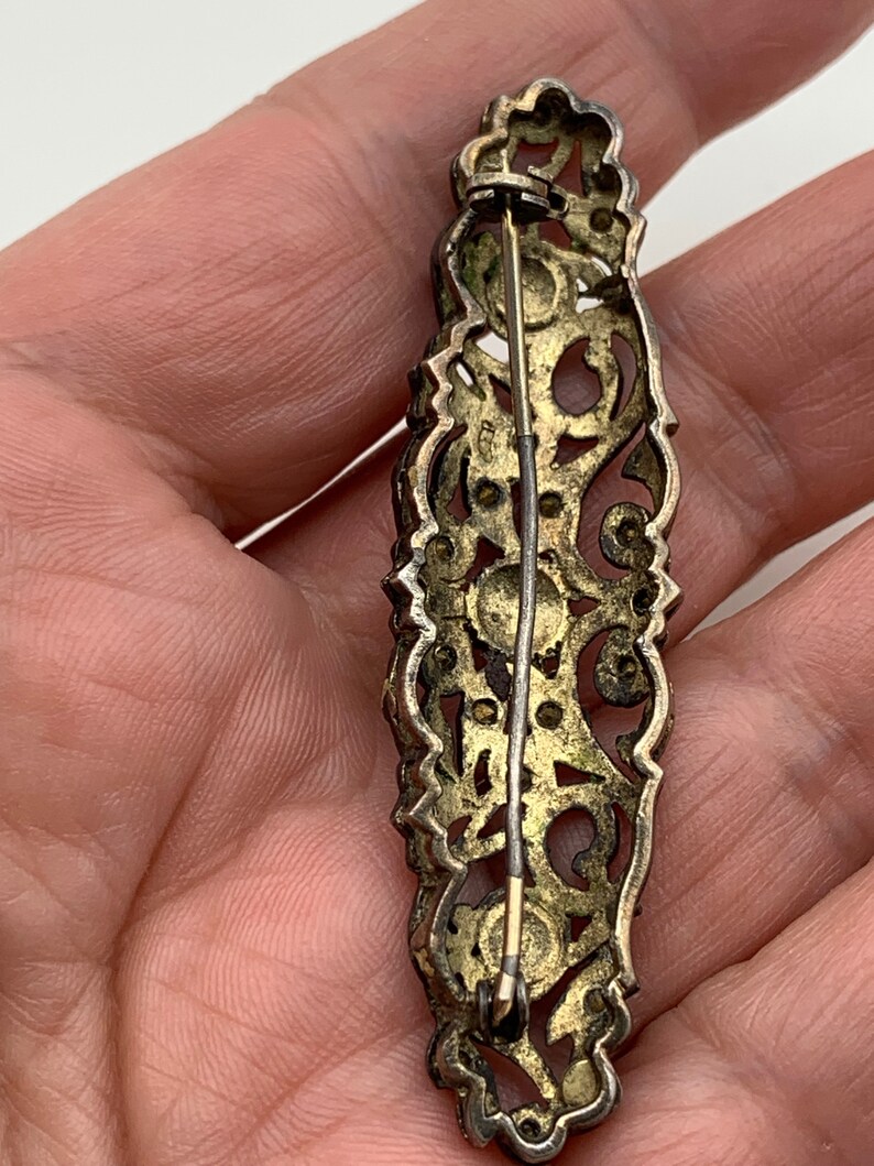 May include: A silver filigree brooch with a gold tone finish and a pin closure. The brooch is decorated with small, round stones.
