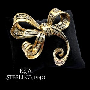 Pode incluir: Um broche dourado em forma de laço com strass transparentes. O broche está marcado com "REJA STERLING, 1940" em texto branco. O design do laço apresenta detalhes intrincados, sobre um fundo preto.