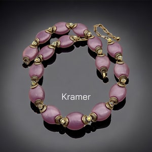 Könnte beinhalten: Eine rosafarbene und goldene Halskette mit ovalen Perlen und kleinen Strasssteinen. Die Halskette liegt auf einem schwarzen Hintergrund und das Wort "Kramer" steht darunter.