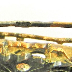 May include: Close-up of a silver brooch with a gold-toned accent. The brooch has a detailed design and the inscription "800" is visible on the back.