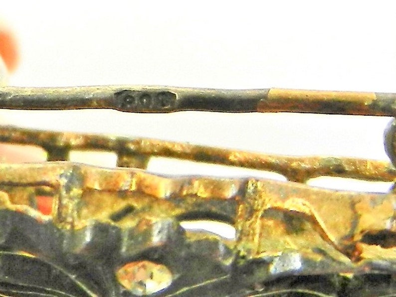 May include: Close-up of a silver brooch with a gold-toned accent. The brooch has a detailed design and the inscription "800" is visible on the back.