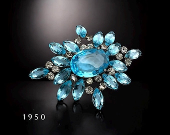 May include: A vintage brooch featuring a large, oval-shaped, light blue rhinestone surrounded by smaller, teardrop-shaped rhinestones in a starburst pattern. The brooch is set in silver tone metal and has a 1950 date stamp.