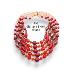 Op de afbeelding: Een statement ketting met meerdere strengen en kralen in tinten lichtroze, dieprood en feloranje. De ketting bevat matte en gefacetteerde kralen in diverse vormen en maten, met een goudkleurige sluiting. Tekst: GM Giuliano Fratti Milano.
