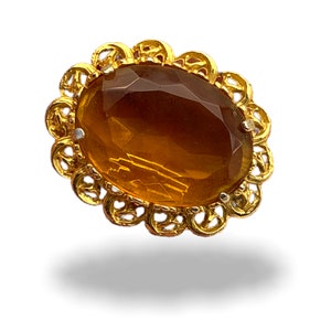 May include: A gold-toned oval brooch with a large, faceted amber-colored glass stone in the center.