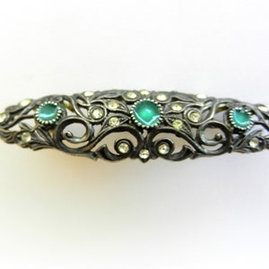 May include: A silver-toned hair clip with a delicate, ornate design featuring green and clear rhinestones.
