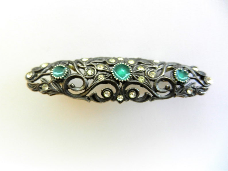 May include: A silver-toned hair clip with a delicate, ornate design featuring green and clear rhinestones.