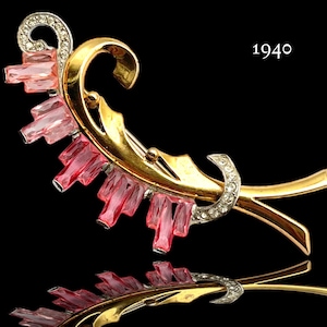 Pode incluir: Um broche em tom dourado e prateado com pedras de vidro rosa. O broche tem a forma de uma flor estilizada com um caule curvo. O ano 1940 está impresso na imagem.