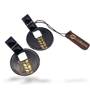 May include: A pair of black and gold earrings with a geometric design. The earrings are round with a square cutout in the centre. The earrings are made by the brand "Laurana".